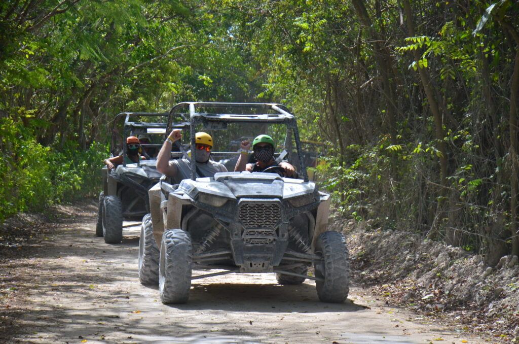 Two muddy side-by-side off-road vehicles with helmeted, masked riders cheering on a shaded forest dirt trail — outdoor off-road adventure.