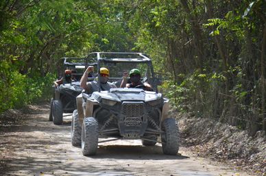 Two muddy side-by-side off-road vehicles with helmeted, masked riders cheering on a shaded forest dirt trail — outdoor off-road adventure.