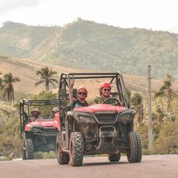 Red off-road UTVs with helmeted riders and a passenger raising a hand, driving on a scenic tropical hill road past palm trees and rolling green hills — outdoor off-road adventure