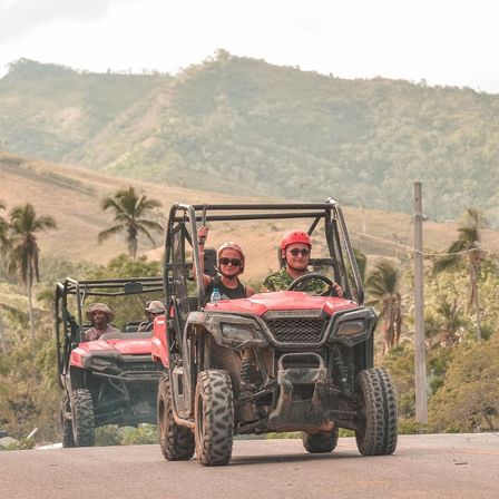 Red off-road UTVs with helmeted riders and a passenger raising a hand, driving on a scenic tropical hill road past palm trees and rolling green hills — outdoor off-road adventure