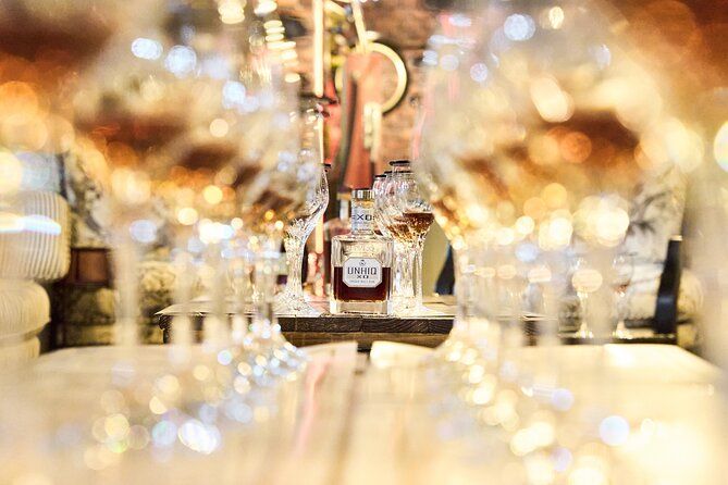 Amber liquor bottle on a wooden tasting table framed by rows of empty wine glasses and warm bokeh lights in a cozy bar or restaurant interior.