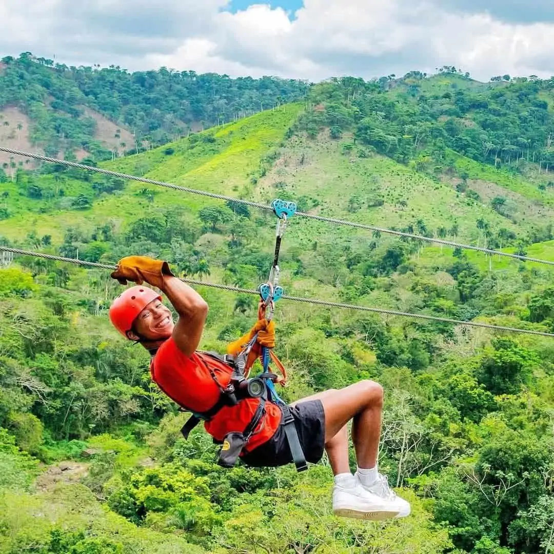 Smiling person in red helmet and shirt ziplining over lush green hills and a tropical forest valley