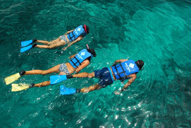 Three snorkelers in blue life jackets and colorful fins floating face-down over clear turquoise tropical ocean water above a coral reef.