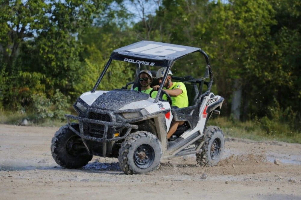 Two people in neon shirts driving a muddy off-road UTV on a dirt trail, kicking up mud near a forested edge — outdoor off-road adventure