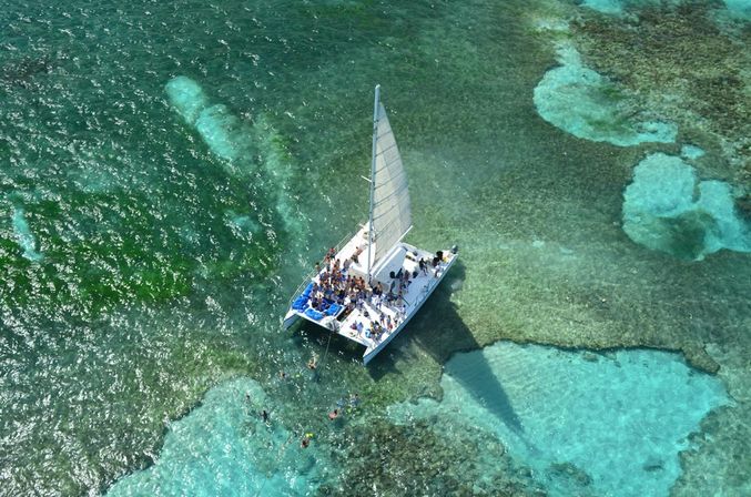 Aerial view of a white catamaran sailboat anchored over a turquoise tropical coral reef, passengers on deck and snorkelers exploring clear shallow waters on a sunny day