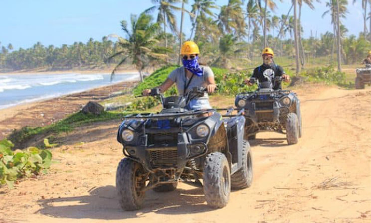 Two riders in yellow helmets driving black ATVs along a palm-lined sandy beach trail with ocean waves and coconut palms under a sunny sky