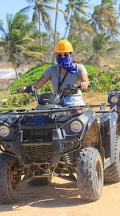 Two riders in yellow helmets driving black ATVs along a palm-lined sandy beach trail with ocean waves and coconut palms under a sunny sky