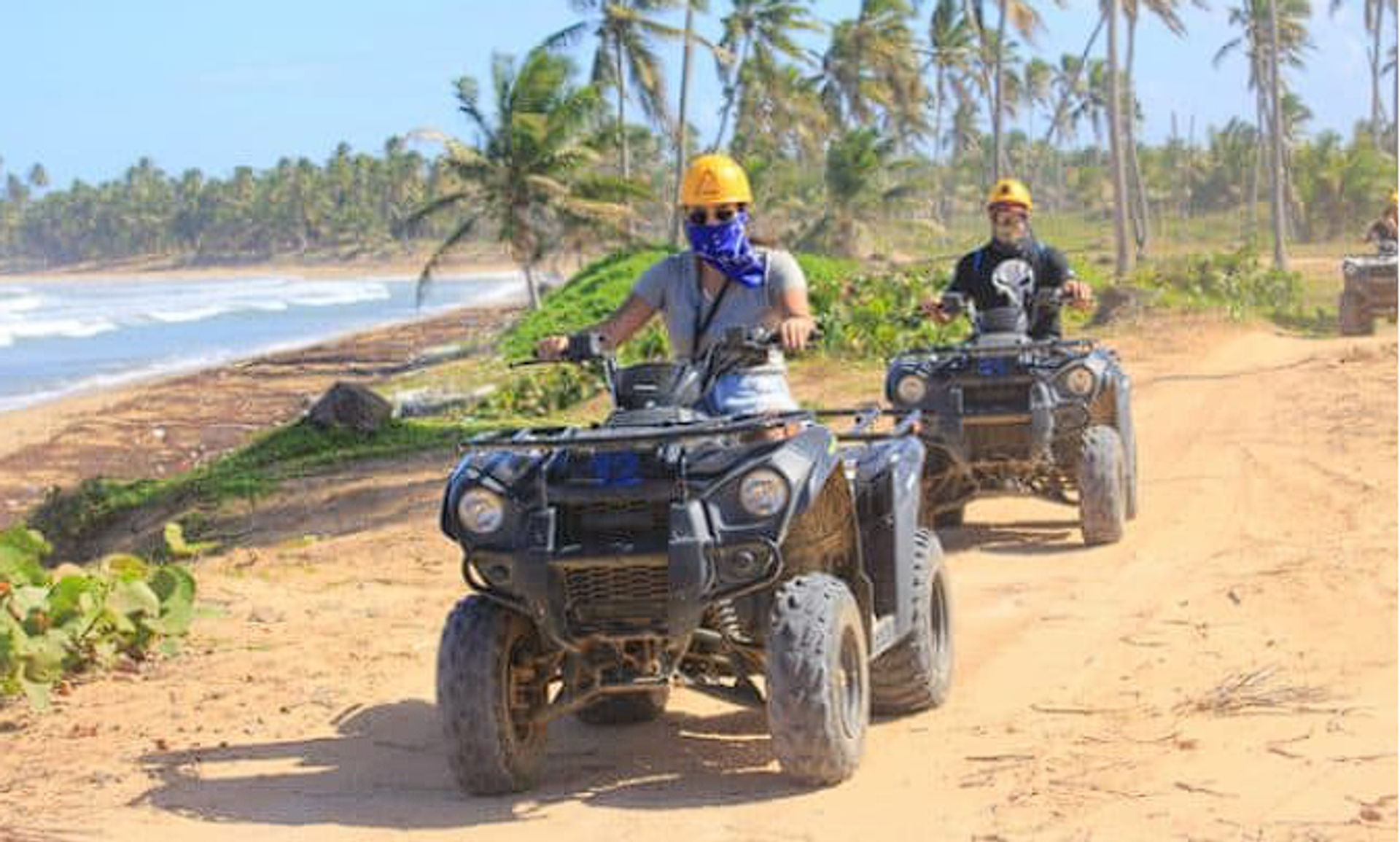Two riders in yellow helmets driving black ATVs along a palm-lined sandy beach trail with ocean waves and coconut palms under a sunny sky