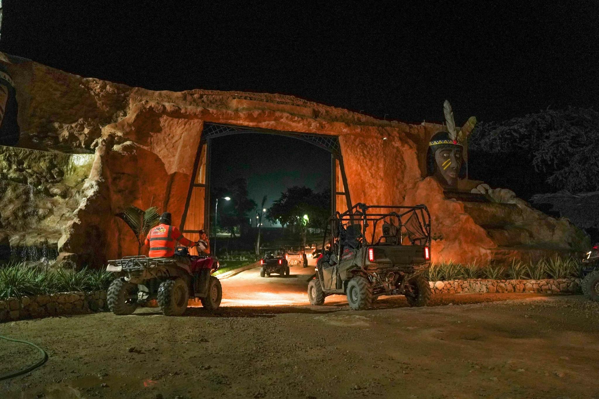 Nighttime adventure scene of mud-splattered ATVs and a UTV passing under an illuminated rock archway flanked by a carved feathered head statue at a park entrance.