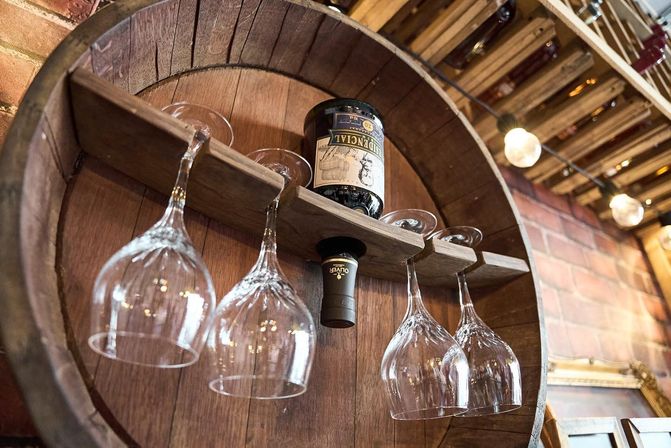 Rustic wooden barrel wine rack mounted on a brick wall holding an inverted wine bottle and four hanging wine glasses under warm Edison bulbs in a cozy tasting room
