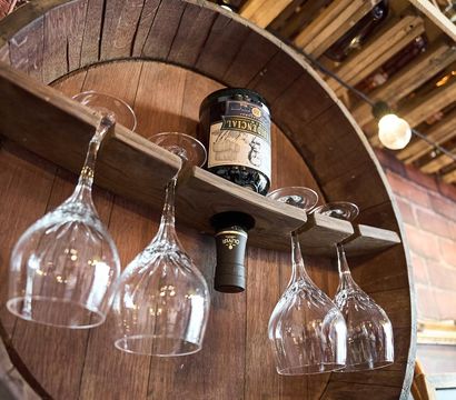 Rustic wooden barrel wine rack mounted on a brick wall holding an inverted wine bottle and four hanging wine glasses under warm Edison bulbs in a cozy tasting room