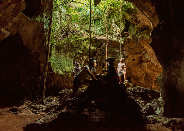 Four helmeted explorers in a sunlit tropical sinkhole cave (cenote-like), standing on limestone rocks beneath hanging vines, mossy walls and an open canopy above — jungle adventure scene.