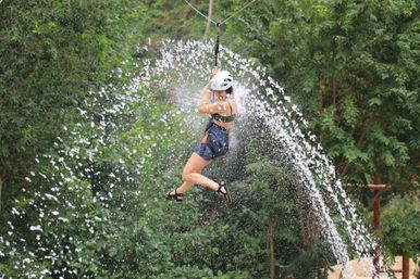 Person ziplining through an arcing wall of water over a lush green forest, wearing a helmet, harness and sandals — outdoor adventure thrill.