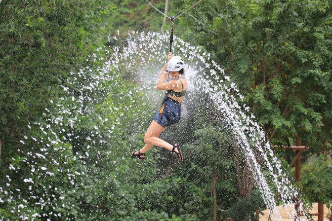 Person ziplining through an arcing wall of water over a lush green forest, wearing a helmet, harness and sandals — outdoor adventure thrill.