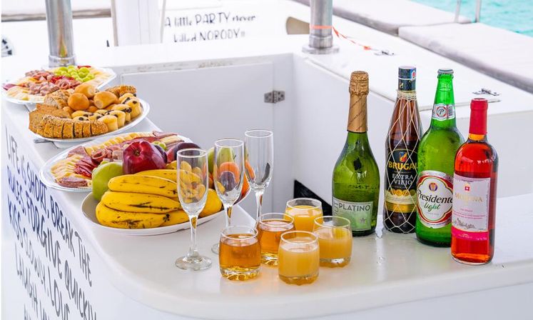 Brunch spread on a white boat deck with platters of bananas, apples, grapes and pastries, champagne flutes and glasses of juice and cocktails, plus assorted wine and beer bottles with turquoise sea in the background.