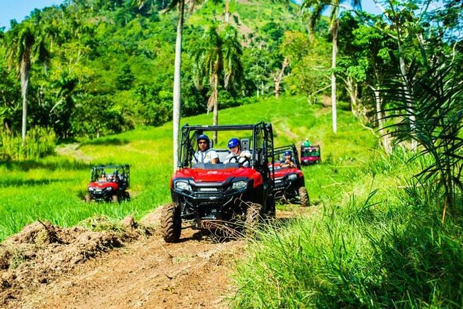 Group of red off-road UTVs on a dirt trail through lush tropical hills and palm trees, riders in helmets enjoying an outdoor adventure