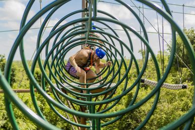 Adult in a blue helmet and safety harness crawling through green circular metal hoops on a treetop ropes course above a leafy forest on a sunny day