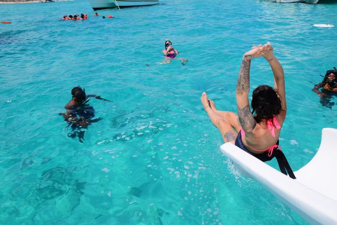 Person with tattoos diving off a white boat into crystal-clear turquoise tropical water, surrounded by snorkelers and anchored boats