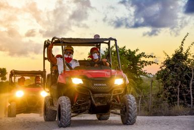 Two red off-road UTVs tearing down a dusty sunset trail, helmeted riders with bandana masks, headlights blazing against tropical shrubs and a colorful sky.