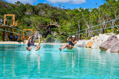 Two riders wearing helmets on parallel ziplines splashing into a turquoise lagoon, with wooden platforms, rocky shoreline and lush tropical forest under a bright blue sky.