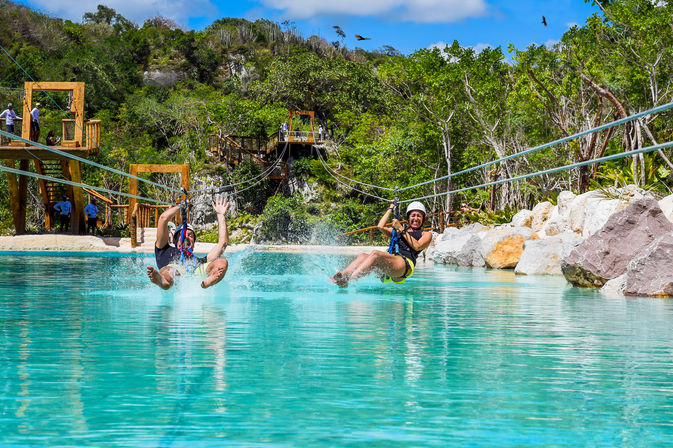 Two riders wearing helmets on parallel ziplines splashing into a turquoise lagoon, with wooden platforms, rocky shoreline and lush tropical forest under a bright blue sky.