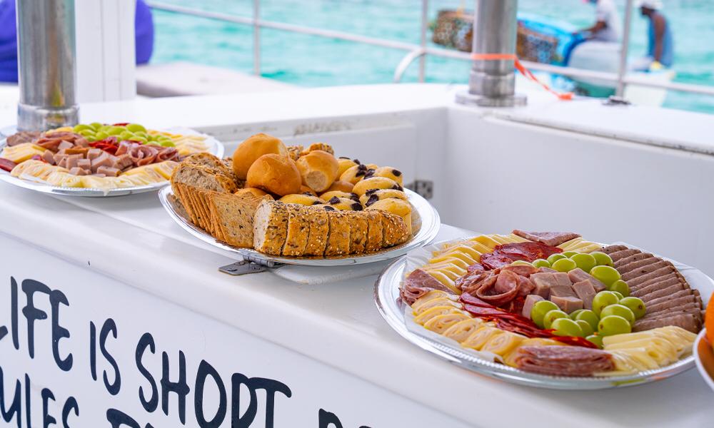 Charcuterie and bread platters with cheeses, cured meats, grapes and rolls displayed on a boat railing against bright turquoise sea.