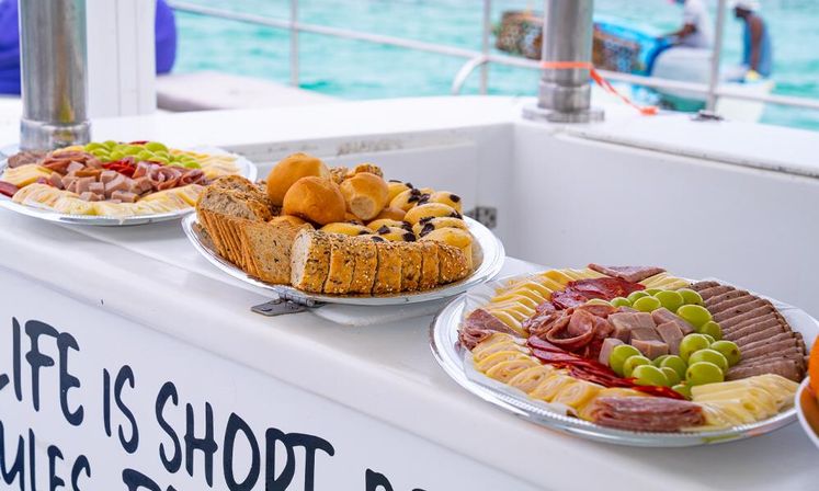 Charcuterie and bread platters with cheeses, cured meats, grapes and rolls displayed on a boat railing against bright turquoise sea.