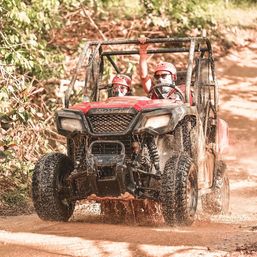 Two people in helmets on a red off‑road UTV splashing through a muddy tropical forest trail — fun off‑road adventure