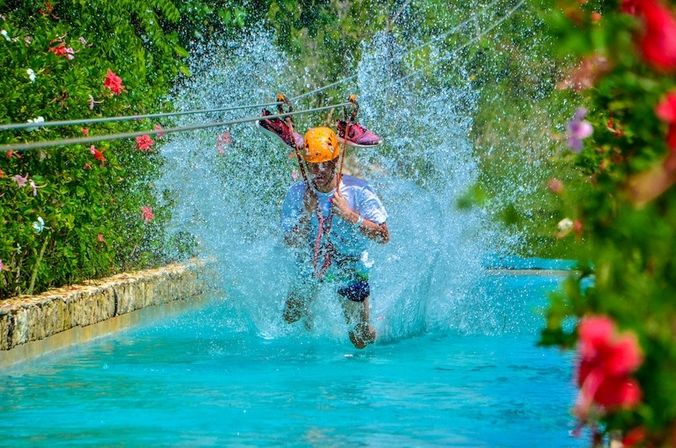 Person in orange helmet and white shirt splashes into a turquoise pool while ziplining through a flower-lined tropical garden.