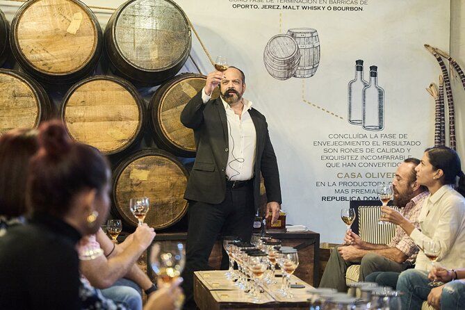 Guided rum tasting in a distillery-style tasting room, presenter raising a glass in front of stacked oak barrels while seated guests sample spirits.