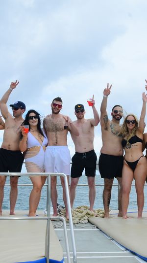 Group of friends in swimsuits raising drinks and peace signs on a boat deck over the ocean, white balloons and a 'LAST DISCO' sign adding a festive coastal party vibe.