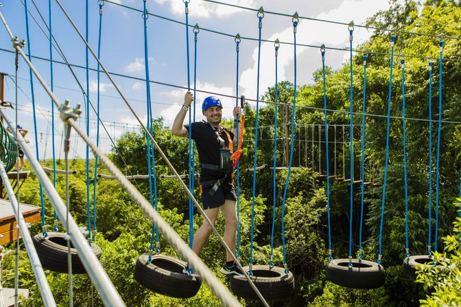 Smiling participant in a blue helmet and safety harness crossing hanging tire obstacles on an elevated outdoor ropes course above a lush green forest canopy — aerial adventure.