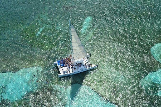Aerial view of a white catamaran anchored over turquoise coral reef with passengers on deck and swimmers/snorkelers in the clear tropical ocean