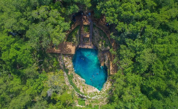Aerial view of a turquoise cenote sinkhole in lush tropical forest, with wooden boardwalks and stairs around the rocky rim and swimmers in clear blue water.