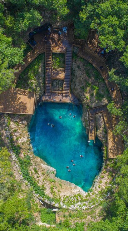 Aerial view of a turquoise cenote sinkhole in lush tropical forest, with wooden boardwalks and stairs around the rocky rim and swimmers in clear blue water.
