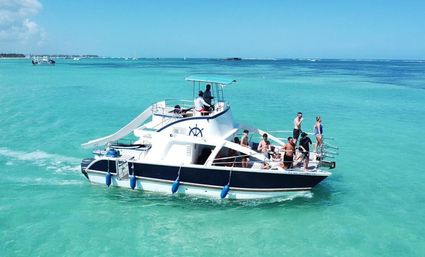 White motor yacht with a water slide and passengers enjoying a sunny boat tour on clear turquoise tropical ocean with a low island coastline in the distance
