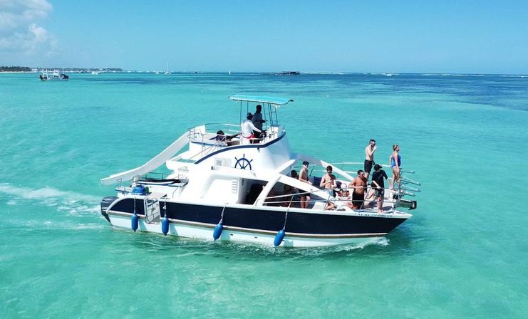 White motor yacht with a water slide and passengers enjoying a sunny boat tour on clear turquoise tropical ocean with a low island coastline in the distance