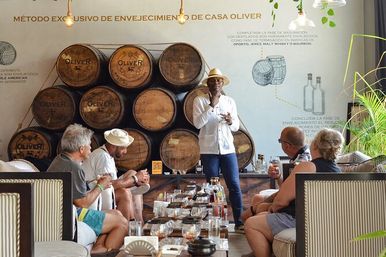 Guide in a straw hat leading a guided rum tasting in a cozy barrel room, guests seated on sofas sampling spirits from bottles and glasses in front of stacked oak barrels.