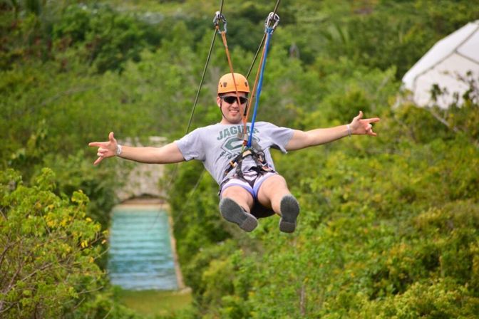 Person wearing a helmet and sunglasses ziplining over lush green tropical foliage toward a turquoise pool, harnessed and arms outstretched in a joyful adventure pose.
