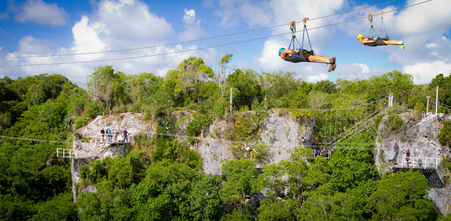 Two riders flying on a cliffside zipline above lush tropical jungle canopy with rocky platforms and a bright blue sky