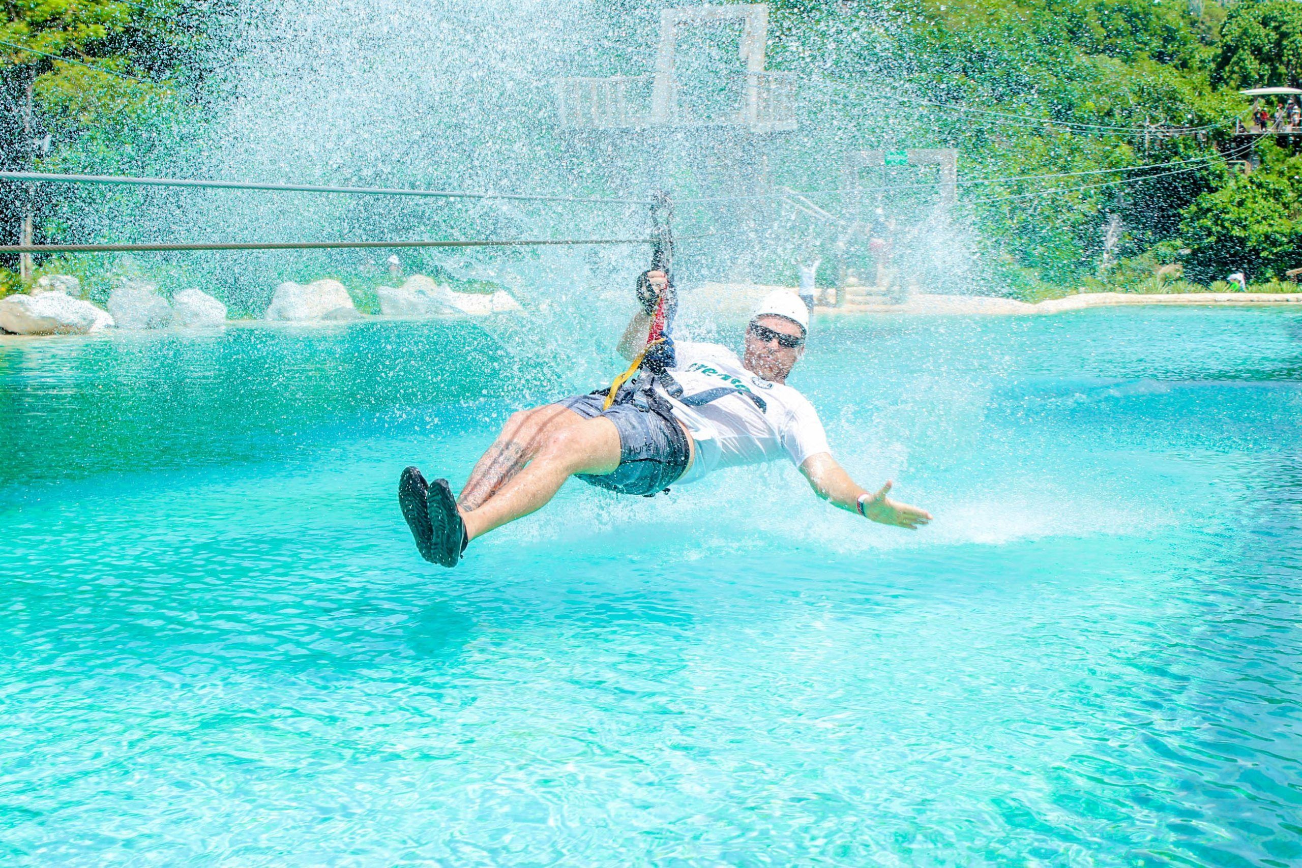 Thrill-seeker on a zipline skims a turquoise pool, sending up a huge splash at a sunny tropical outdoor water park, wearing a helmet and harness.