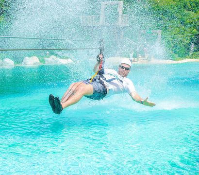 Thrill-seeker on a zipline skims a turquoise pool, sending up a huge splash at a sunny tropical outdoor water park, wearing a helmet and harness.