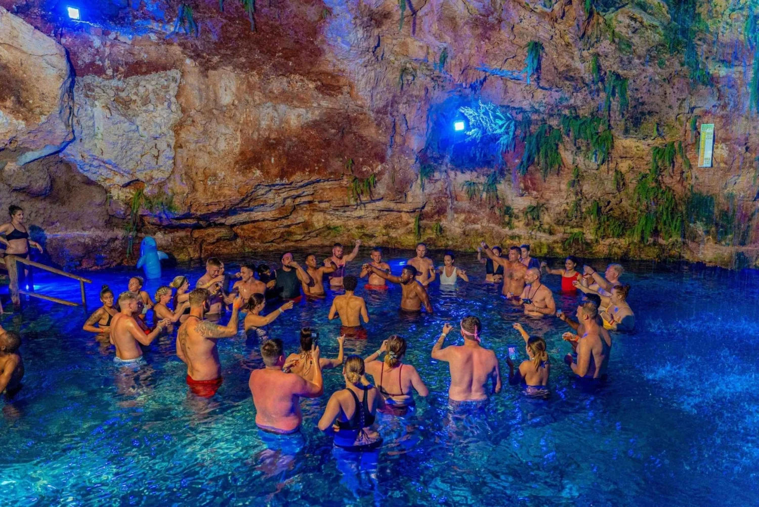Lively group of swimmers and tourists gathered in a blue-lit underground cenote cave pool, wading in clear water near rocky walls and hanging greenery.