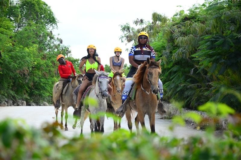 Group of four riders in helmets on a guided horseback riding tour along a sunlit tropical trail surrounded by dense green foliage