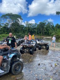 ATV tour group parked on a rocky riverbank in a lush tropical rainforest, riders chatting by shallow water under a bright blue sky with puffy clouds.