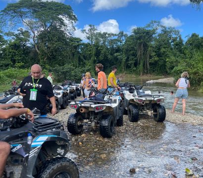 ATV tour group parked on a rocky riverbank in a lush tropical rainforest, riders chatting by shallow water under a bright blue sky with puffy clouds.