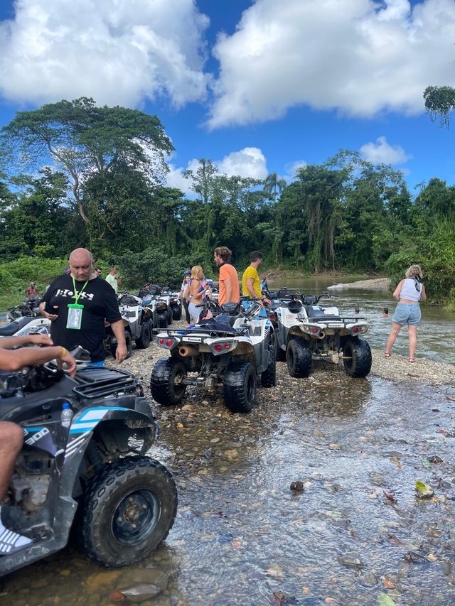 ATV tour group parked on a rocky riverbank in a lush tropical rainforest, riders chatting by shallow water under a bright blue sky with puffy clouds.