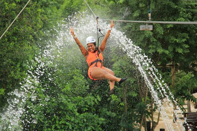 Thrilled person in orange harness and helmet ziplining through an arching spray of water over lush tropical greenery, arms raised flashing peace signs.