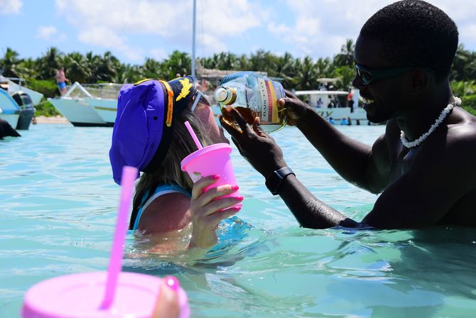 Playful tropical beach scene in shallow turquoise water: a man pours rum from a bottle into a woman’s mouth as she holds a pink cup and wears a purple captain hat, with boats and palm trees in the background.