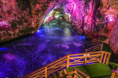 Glowing underground cave grotto with vibrant purple and blue lights reflecting on a clear subterranean pool, wooden stairs and railings forming a stepped viewing platform down to the water.